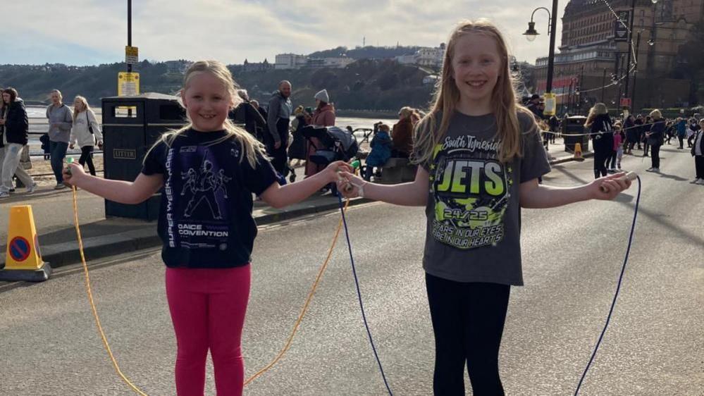 Two girls stand smiling on the Scarborough Foreshore holding skipping ropes. 