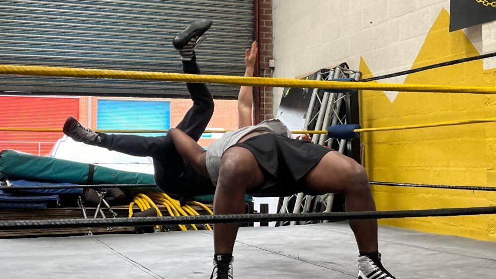 The character Lysander preparing to slam the character Demetrius to the ground inside a wrestling ring.