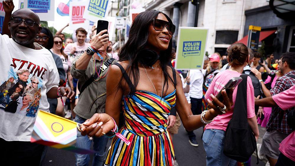 Naomi Campbell wearing a multi-coloured striped dress stands with groups of parade participants. She is wearing sunglasses. 