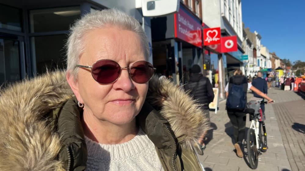 Dawn Henney, wearing prescription sunglasses, and a knitted Arran style jersey and a green Parka jacket with faux fur lined hood. She is standing in Great Yarmouth market place where other people are passing by on foot or with a bicycle.