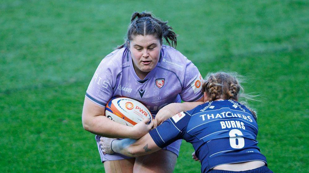 Maud Muir runs into a tackle from Bristol's Delaney Burns, holding a rugby ball close to her body under her right arm during a game for Gloucester-Hartpury this season