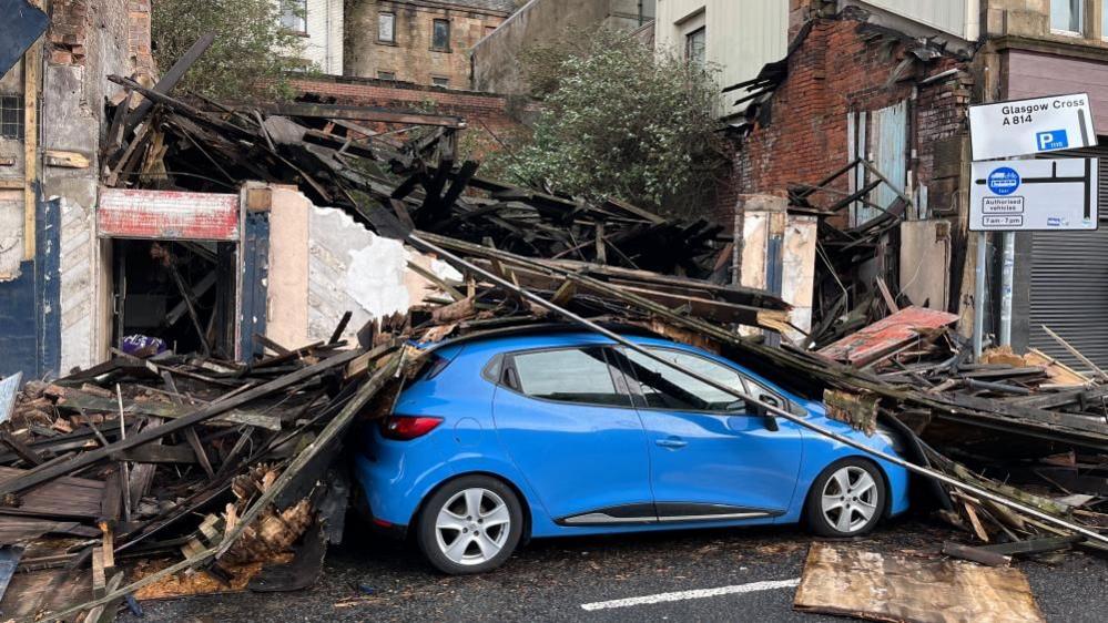 A small blue car lies buried underneath pieces of wood from a collapsed building behind it.