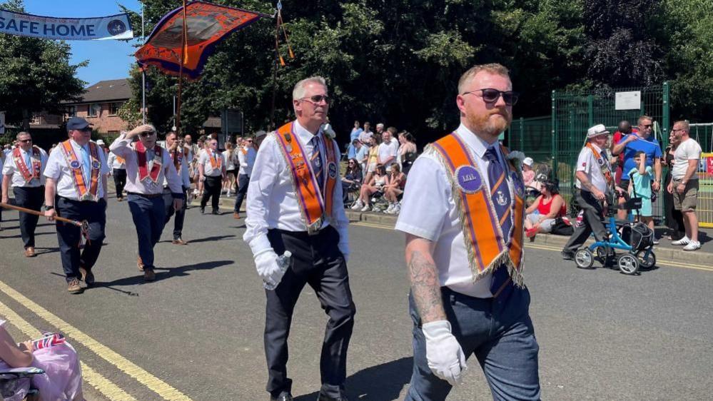 Men walking in the parade wearing orange sashes and white shirts.