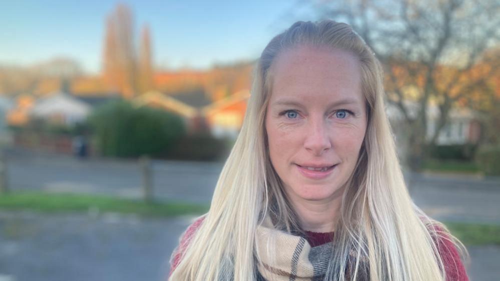 A woman with long blond hair, in a checked scarf and red coach. She is stood on a pavement in the centre of a village.