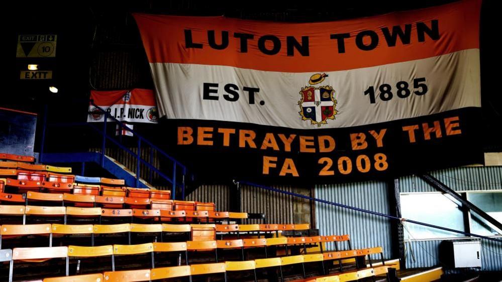 A Luton Town flag by the side of a stand at Kenilworth Road, Luton, that says "Luton Town Est. 1885 Betrayed by the FA 2008"