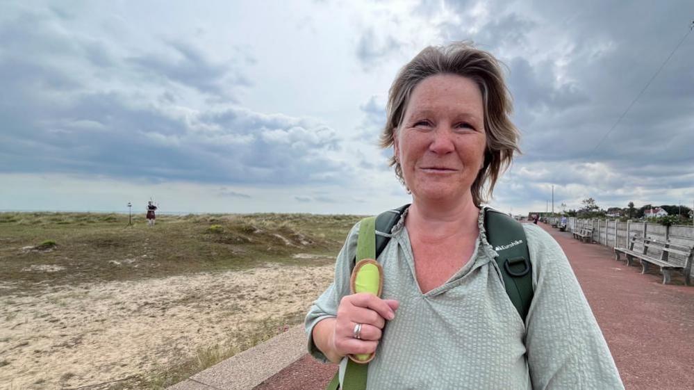 Ellen Bygrave medium length straw blonde hair, and is wearing a green v-neck top, with green rucksack straps over each shoulder and a lime green bag strap over her right shoulder. She is smiling, standing on the promenade of Great Yarmouth's north beach. Piper Ryan Fitzpatrick is standing on the grass dunes in the distance. Benches and fencing are lining the right side of the promenade.
