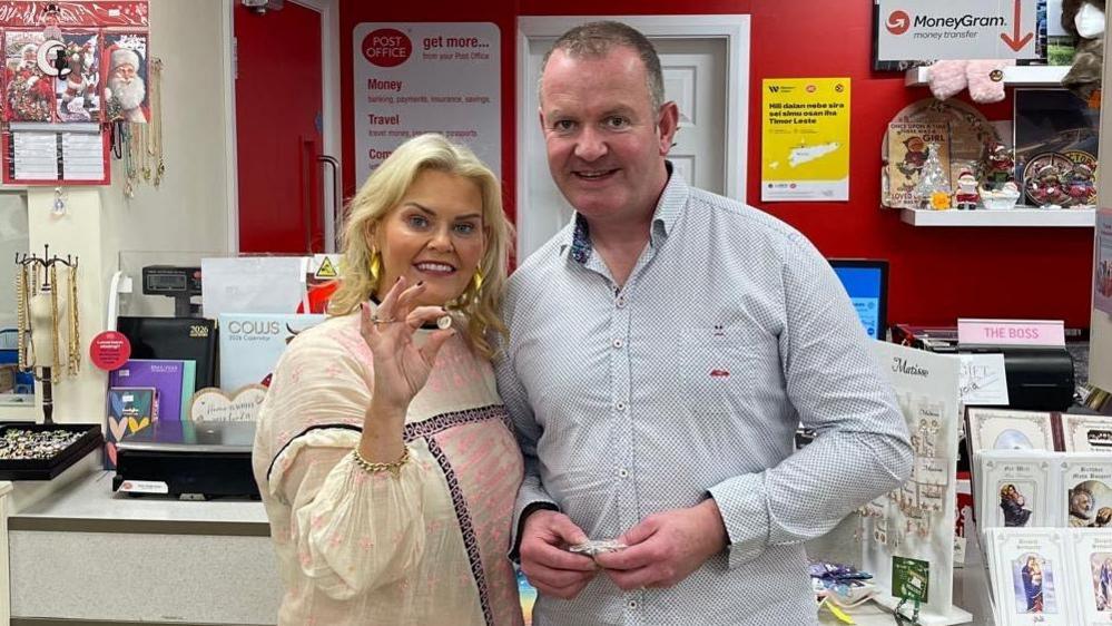 Sean Ferry standing next to his wife Olga. Sean has short shark hair, wearing a light blue shirt. Olga has shoulder-length blonde hair, wearing a cream top. She is holding up the 5p coin in her right hand.