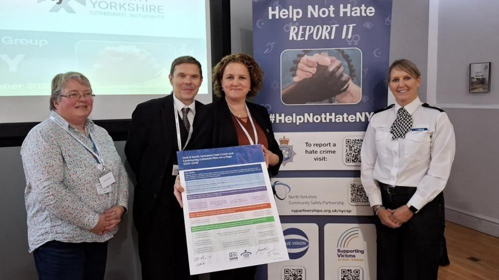 Four people stand in front of a banner reading "help not hate - report it" - holding a large piece of card with colourful blocks