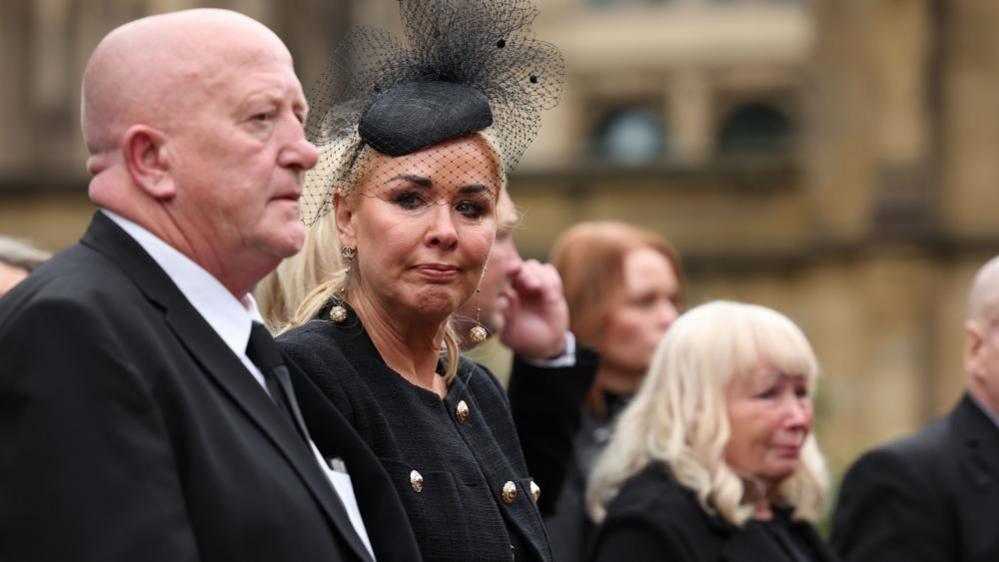 Clare Sweeney stands wearing a black jacket and veiled headband. She has teas in her eyes.