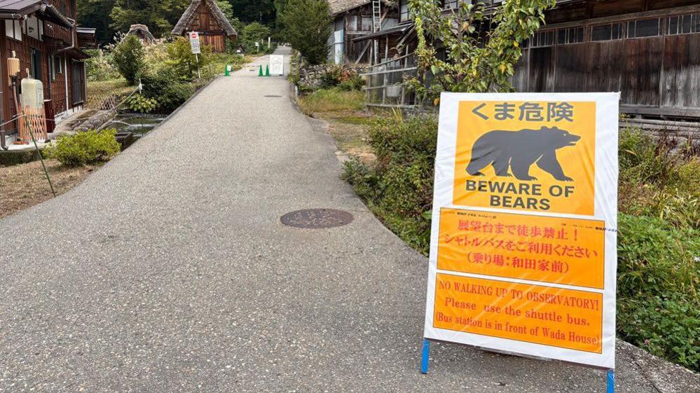 A yellow sign propped on a road that reads: Beware of bears. In the background are traditional Japanese wooden houses.