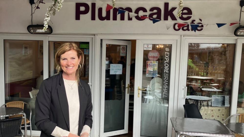 A woman with long blond hair stands in front of a shop front which reads Plumcake Cafe. 