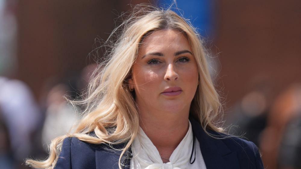 Ms Hand has long blonde hair. She has a plain expression on her face. She is wearing a white blouse and navy jacket.