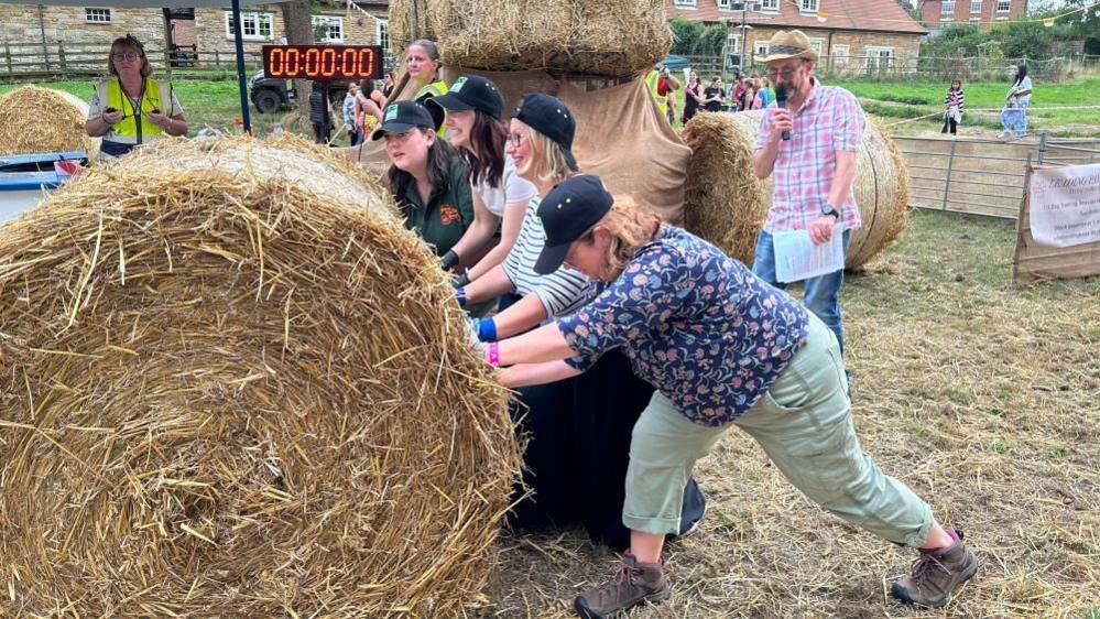 A group of women in blue caps push a large straw bale around a grass course. A digital timer is visible.  A man with a microphone is commentating.