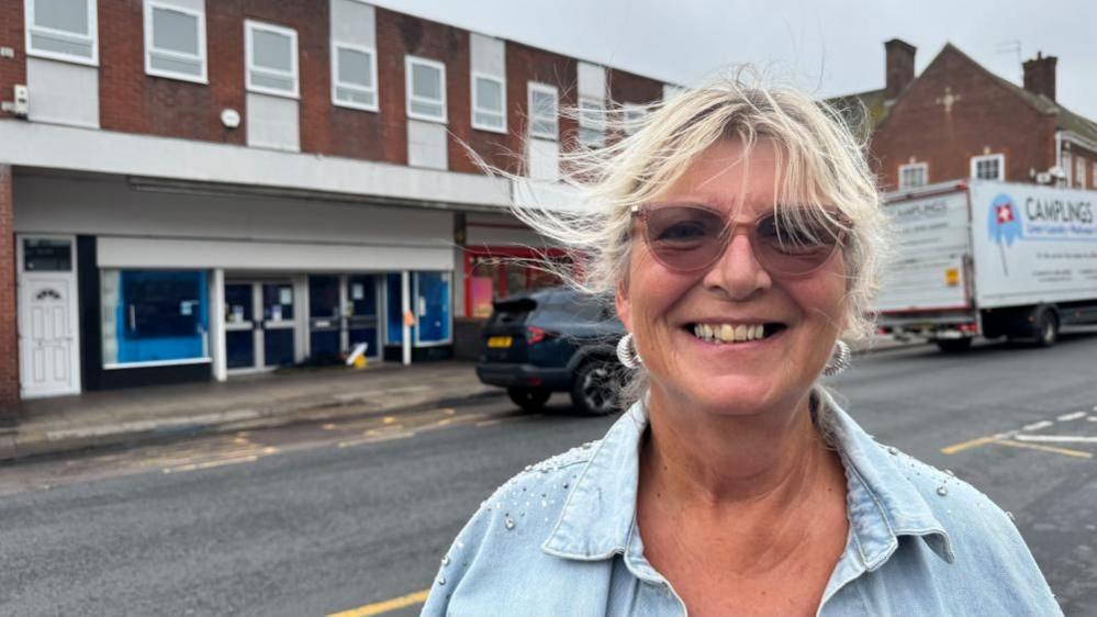 Maureen Paterson is wearing a light denim shirt with studs and diamante decoration on the shoulders. She is wearing earrings, and tinted glasses. She has blonde hair, and is smiling.