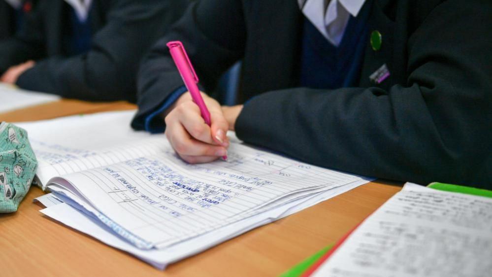 A generic image of a pupil writing in an exercise book. It is a close up showing the desk, with a lined book on it with paragraphs of text. The pupil is poised as if writing. The pen is pink. You can see their arms and up to their collar, although the background is slightly blurred.