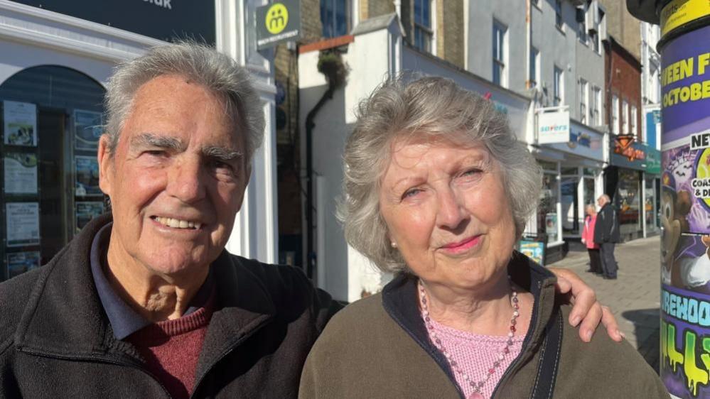 David Cowling stands beside his wife Frances Cowling, 79. They are both looking directly at the camera and smiling. They both have grey hair. He is wearing a black fleece jacket, blue collared polo shirt and a red jersey. She is wearing a taupe fleece top, with a pink necklace and pink knitted top. He has his hand on her left shoulder. They photographed outside on a shopping street. 