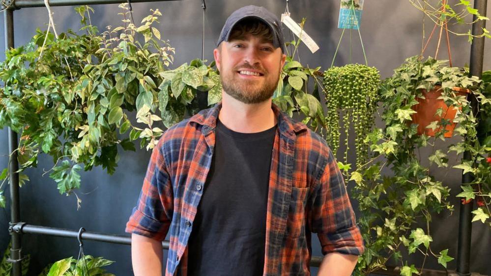 Tony Le-Britton, standing in his shop, Not Another Jungle, by plants, wearing a checked shirt, a blue cap and smiling to the camera