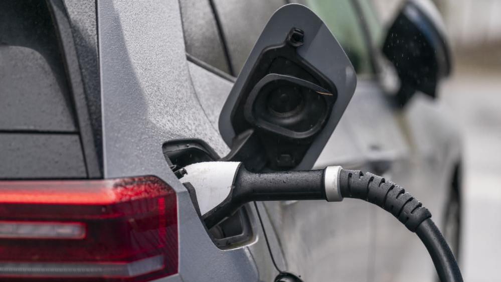 An electric car is plugged in charging. The back right corner is visible, with the charging cap open and a charging cable plugged in to the side of the car. The car itself is grey and is covered with raindrops.