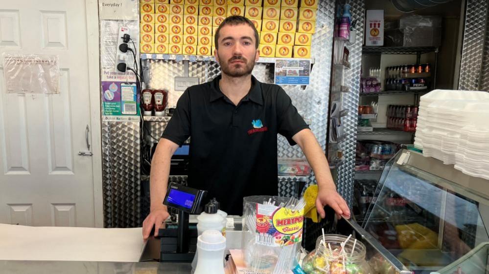 Omer Aydin with short dark hair and beard wearing a black T-shirt behind the counter of a chip shop. Battered fish is visible in the heated display unit.  Salt and vinegar is also visible, along with lollipops.