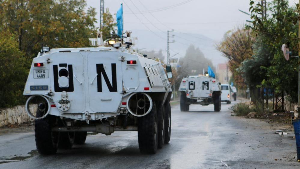 Two white armoured vehicles drive along a road in Lebanon, with UN painted on the backs and the blue flag flying atop.