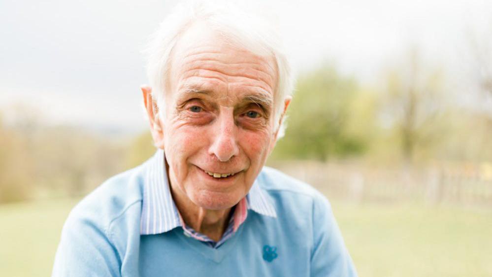 Mr Standring smiles at the camera in a posed photo in a park. He wears a light blue jumper and a pink, white and blue striped shirt, and has grey hair.