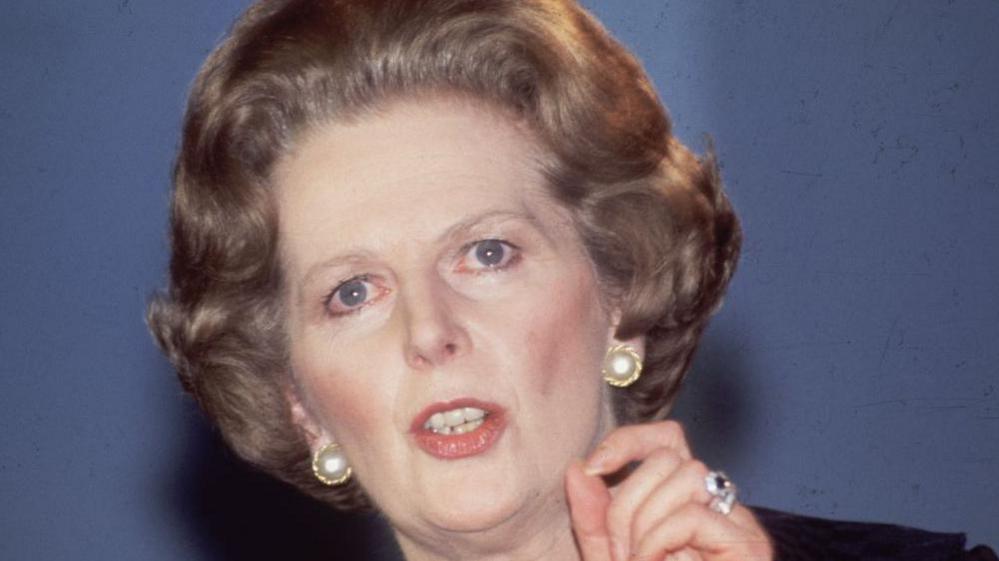 Archive photograph of Margaret Thatcher taken in 1984. She has dark-blonde hair and blue eyes, is wearing pearl-stud earrings and is holding her thumb and forefinger together as she makes a speech. On her third finger is a jewelled ring.