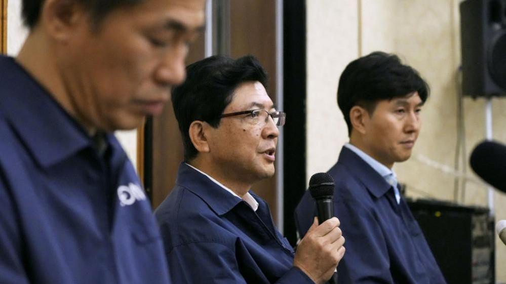 Aeon President Akio Yoshida speaking with a microphone between two other men
