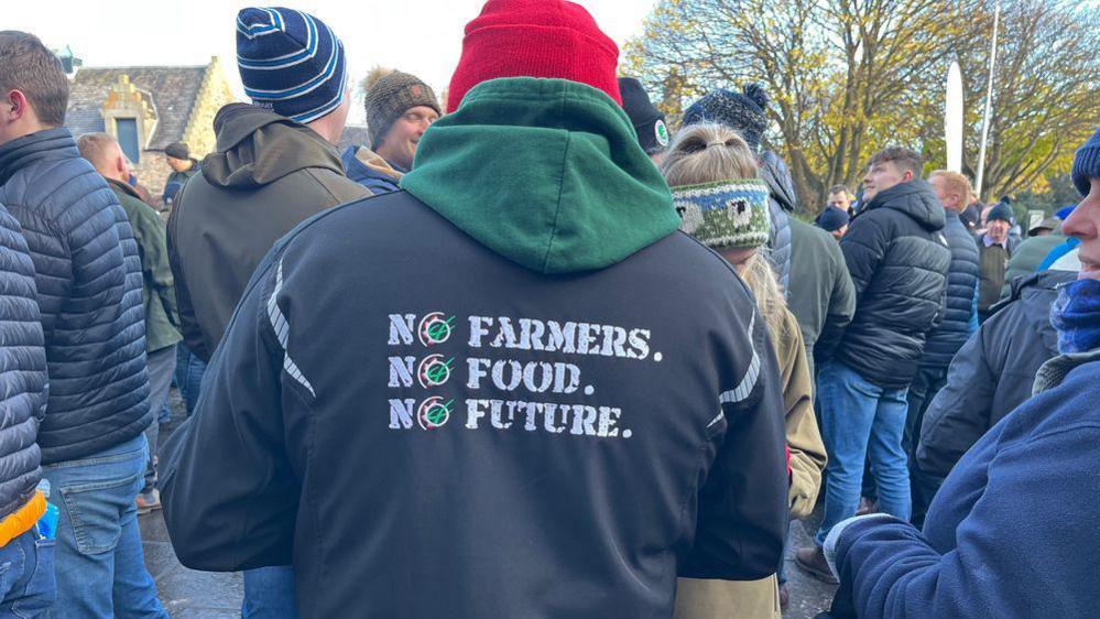 A man wears a jacket and the camera shows the back which reads "No farmers, no food, no future". A crowd is in the background