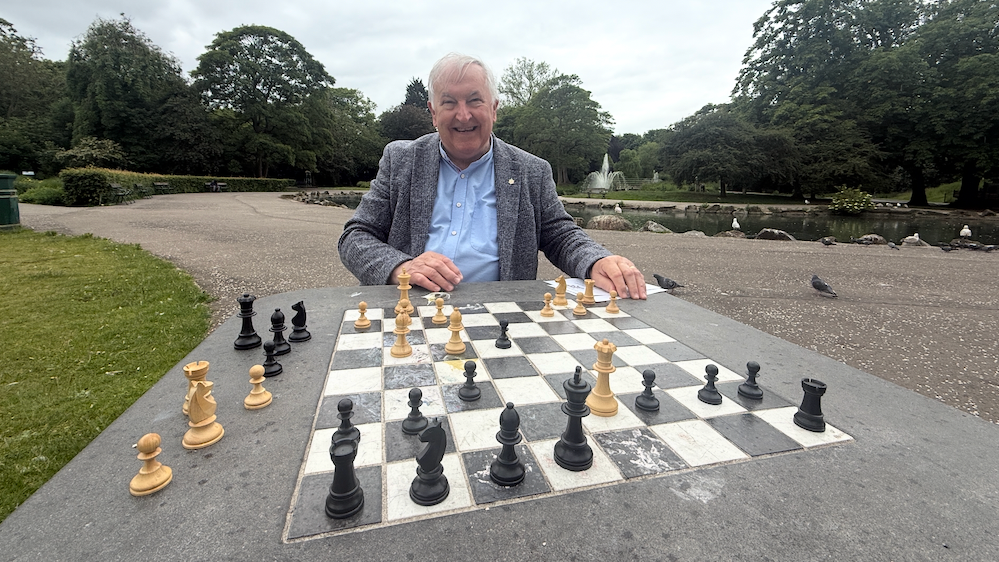 Graham Chesters has white hair and is wearing a grey jacket with a blue shirt underneath. He is sat down at a concrete chess board in a park near a pond. The black and white chess pieces are scattered around the board. He has just achieved checkmate with his queen and bishop angled towards the opposing black king.