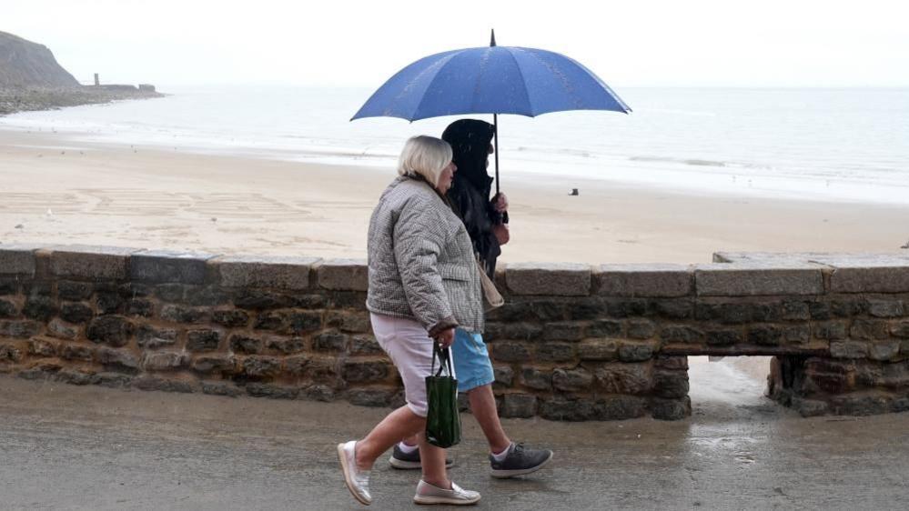 Two women walk along a beach promenade clutching an umbrella.