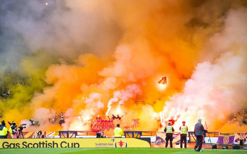 Motherwell fans hold up bright claret and amber pyrotechnics during a Premier Sports Cup semi-final match between Motherwell and Rangers at Hampden Park in 2024