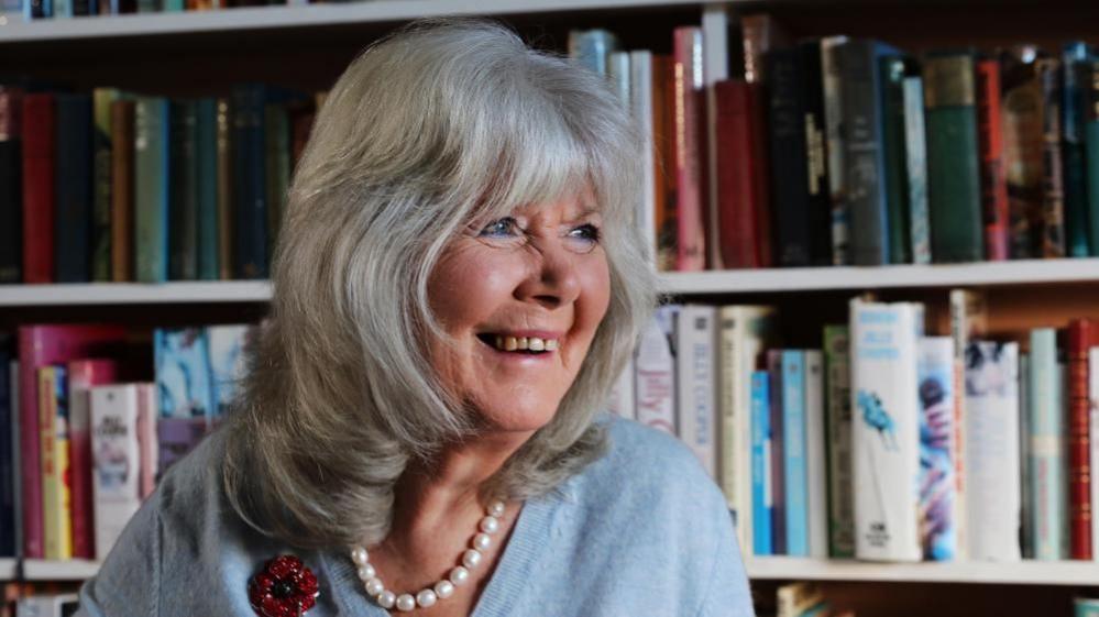 Best-selling author, Jilly Cooper, in front of a packed bookshelf at her home in the village of Bisley in Gloucestershire. Jilly is wearing pearls, a blue jumper and a sparkly Poppy. She smiles as she looks beyond the camera to the right. 