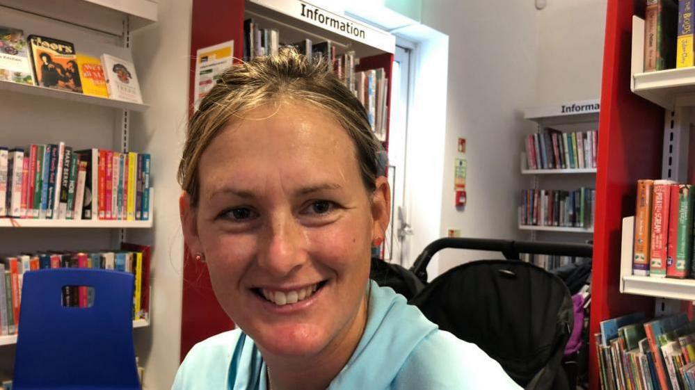 A woman with blonde hair is sitting in a library with a pushchair in the background