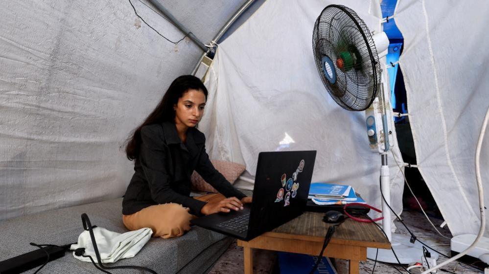 Palestinian student Raghad Loai Mhanna, who passed her high school exams while living in a tent after being displaced during the war, works on her laptop inside her tent in Deir al-Balah, central Gaza Strip on 19 October 2025.