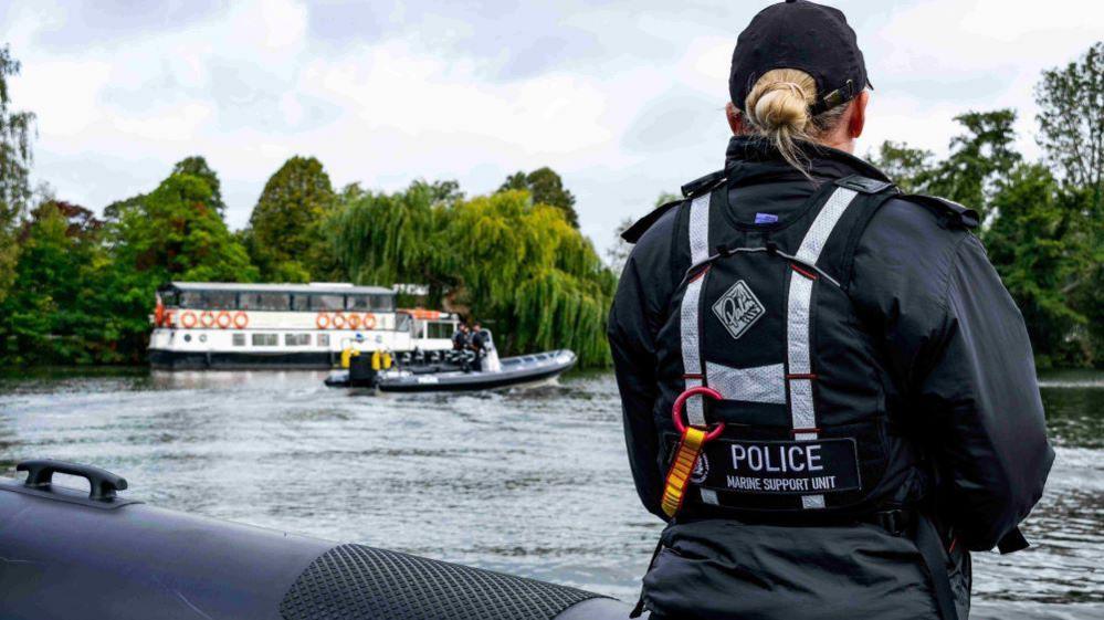 A female marine support unit officer standing on a rib boat looking across the river with her back to the camera.