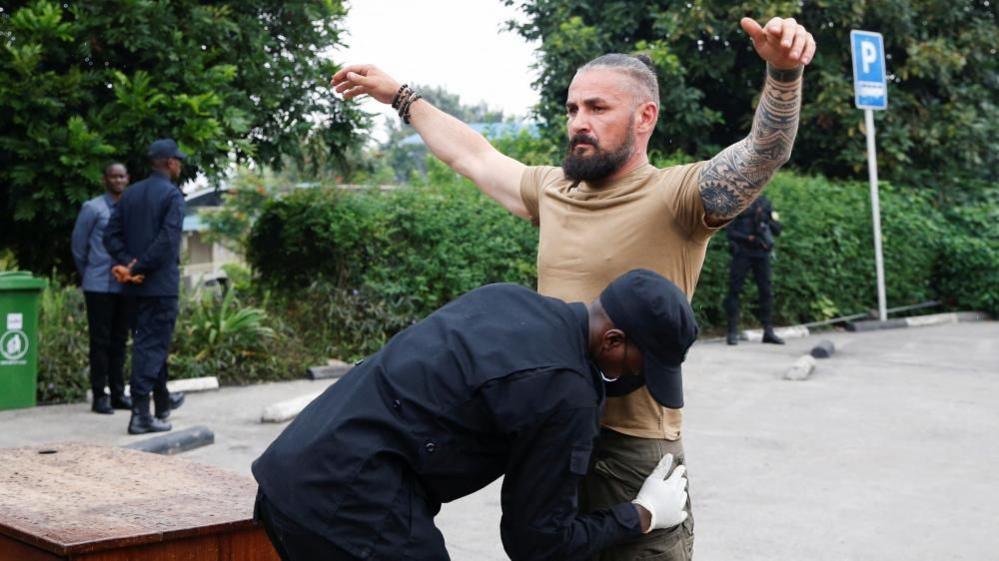 A man in a T-shirt has his tattooed hands raised as he is searched by a Rwandan officer