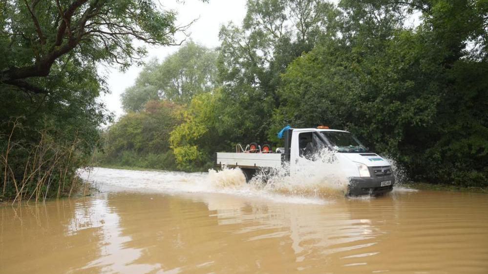 A white lorry containing an upside-down wheelbarrow is moving through brown floodwater and creating a spray and wash. There are trees either side of the road.