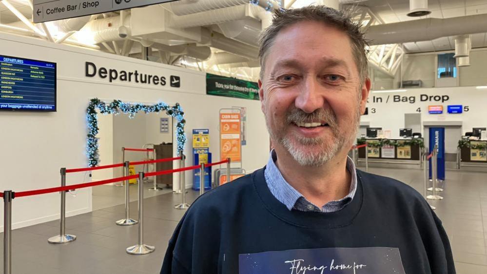 Steve is smiling at the camera and has short brown/grey hair. He has a greyish goatee style beard. He is wearing a navy jumper with a blue collar of a shirt poking out of the top. 