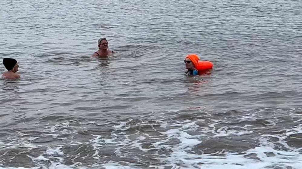 Pam Spychal, with an orange hat and float, swimming in the sea with two of her friends, to raise money for charity. Pam is on the right of the image.