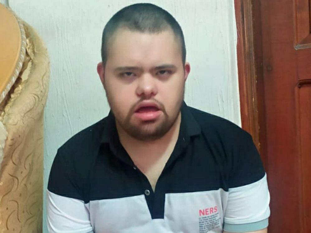 Muhammed, a young man with Down's Syndrome looks at the the camera. He has dark hair which is cut very short and a short beard. He is wearing a polo t-shirt which is black and white. He is sitting in a room next to a sofa, with a painted wall and a dark wooden door behind him. 