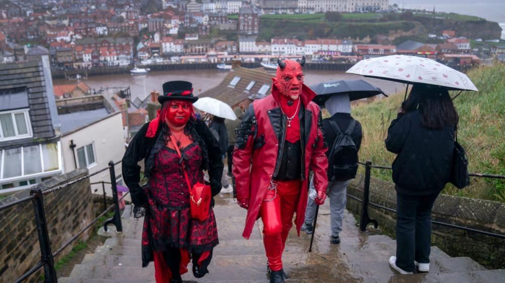 A man and a woman in red face paint and gothic clothing walk up Whitby steps