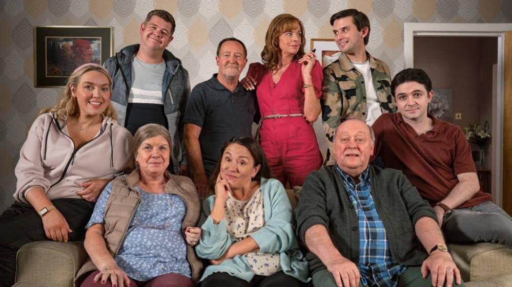 A group of eight people gathered in a living room setting. Four individuals are seated on a couch in the foreground, while four others stand behind them. The background features patterned wallpaper with a leaf design in muted tones.