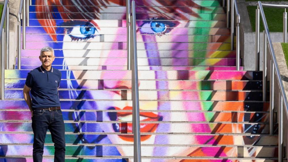Mayor of London Sir Sadiq Khan stands on steps outside Wembley Stadium, which have been painted with a multicoloured mural of Taylor Swift's face. Sir Sadiq is stood on the left hand side, and is wearing a dark blue polo shirt and dark blue jeans.