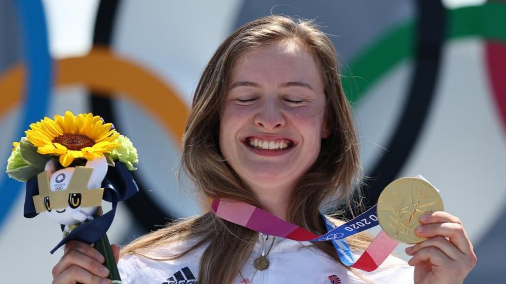 Worthington with long brown hair wearing a white Team GB jacket and holding a sunflower in one hand and a gold medal in the other. She is smiling and has her eyes closed. The Olympic rings are visible behind.