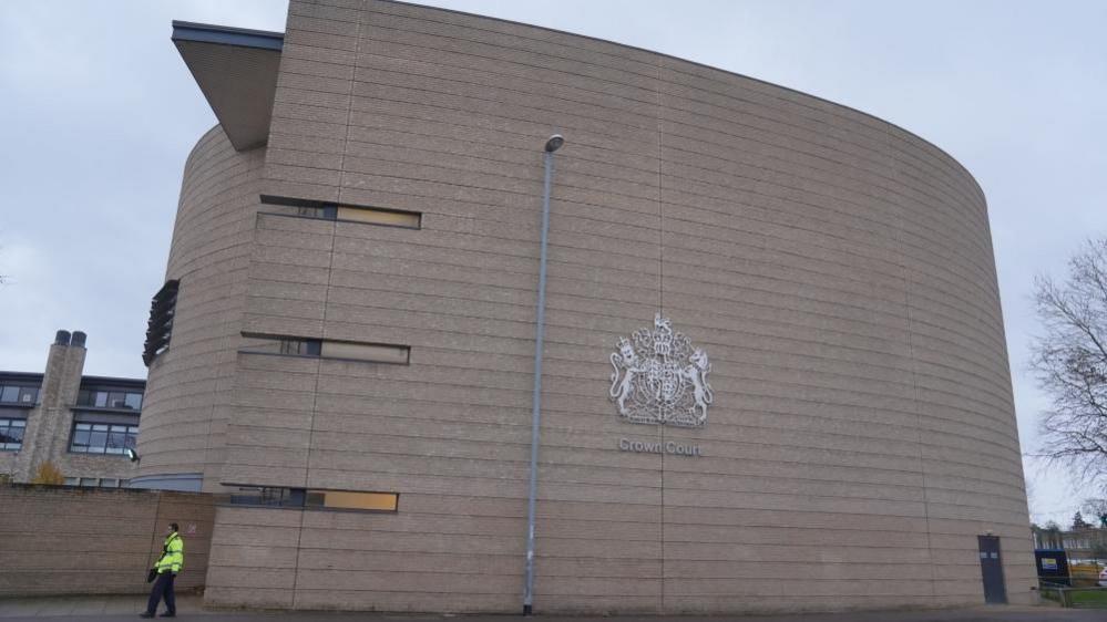 Cambridge Crown Crown: A grey building marked with a crest and words "Crown Court". A figure wearing a yellow jacket and dark trousers is walking in the bottom left of the image.