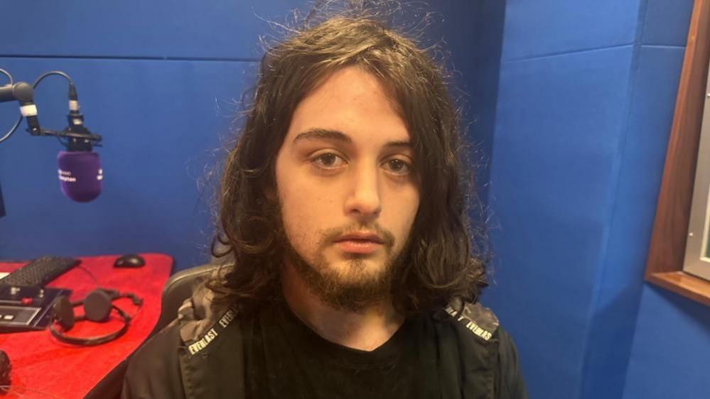 A young man with long black hair and a beard looks at the camera. He is wearing a black T-shirt and jacket and is sat in a radio studio.