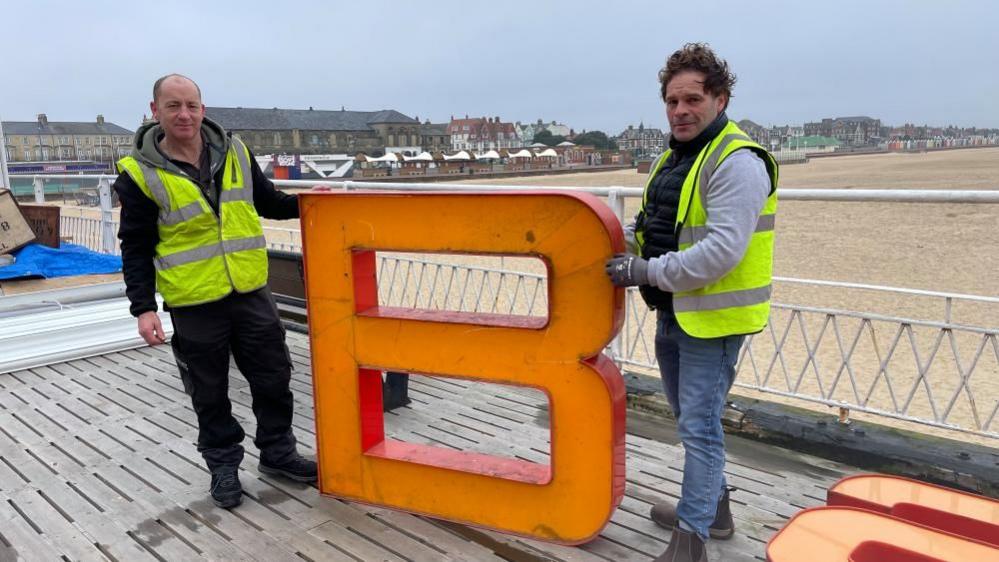 Memorabilia collector Kaine Roo standing with his friend who helped him collect the Britannia Pier lettering. They are holding the letter 'B' and behind them is the beach and seafront buildings including the Arc Cinema. Both men are wearing high visibility jackets and workwear clothing.