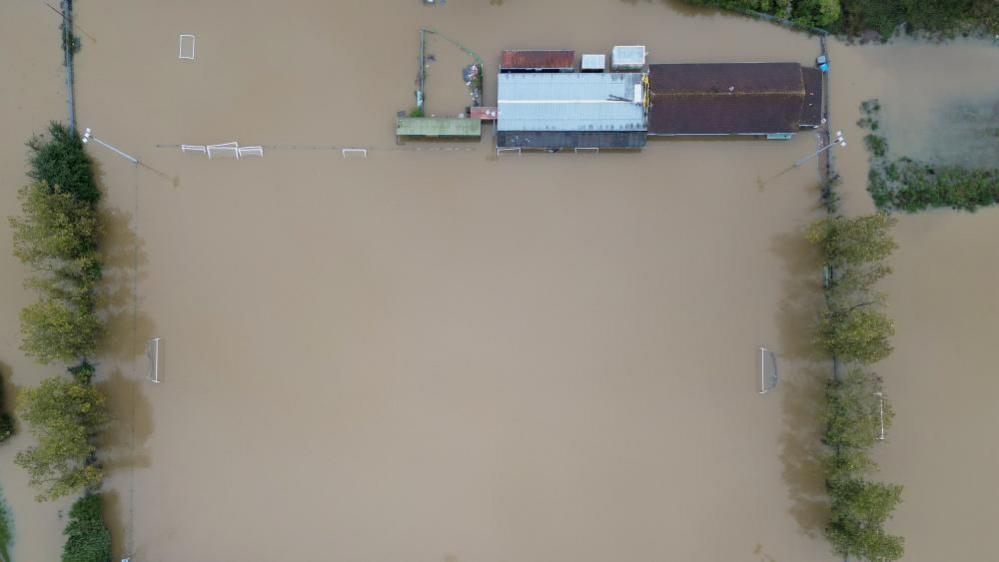 Floodwater in which football goals can be seen from the air submerged. There is a club house to the top right with a white roof and another building with a brown roof.  Trees flank the pitches on either side.