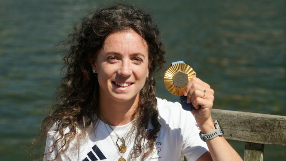 Imogen Grant, holding a gold medal in her left hand, looking at the camera, wearing a Team BH white top