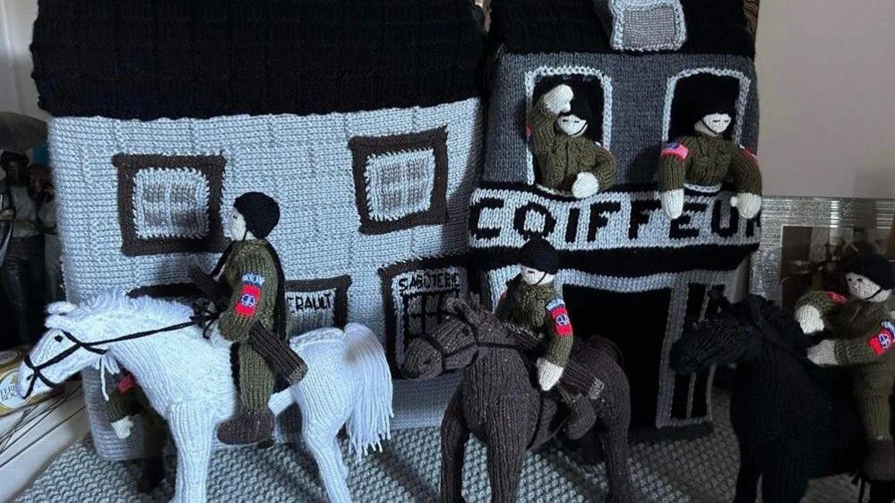 A knitted scene featuring black and white houses with soldiers peering out of the windows. There are three horses standing in front of the houses, a white, brown and black horse. Each horse has a soldier sitting on top of them 